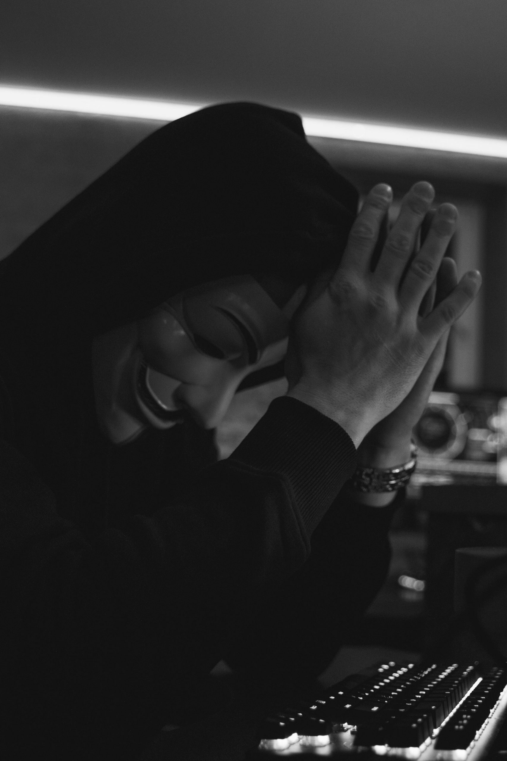 A man wearing a Guy Fawkes mask in a black hoodie sits indoors, symbolizing cyber security and hacking.